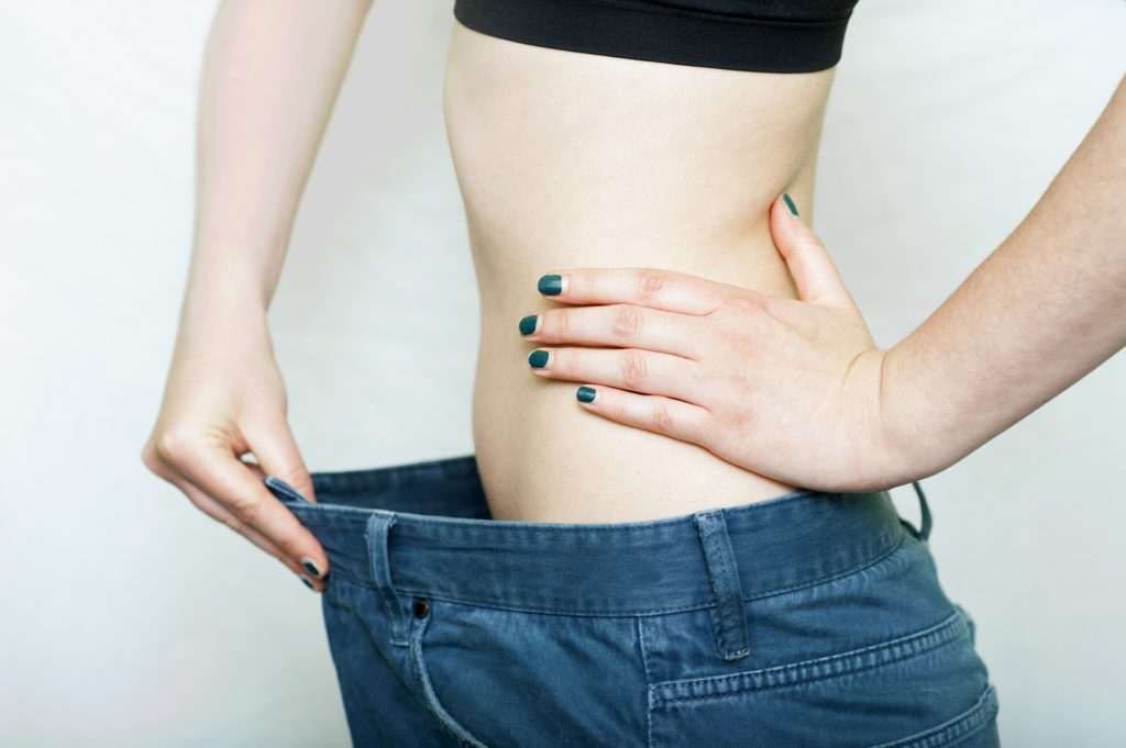 Photograph of keto dieter holding out their pants waistband used to illustrate, "5 Keto Tips For Getting Rid Of Stubborn Fat". Click to learn about getting a customized keto diet plan designed based on your activity level, food preferences, weight goals, and other personal criteria.